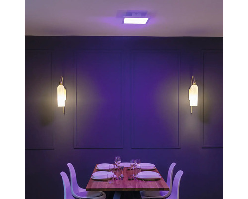 Modernes Esszimmer mit quadratischem LED-Deckenpanel in Lila und zwei Wandleuchten vor einer dunklen Wand mit gedecktem Holztisch und weißen Stühlen.