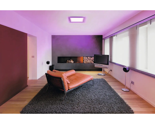Modernes Wohnzimmer mit quadratischem LED-Panel an der Decke, das violettes Licht spendet, orangefarbene Liege auf grauem Teppich.