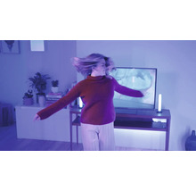 Eine Frau tanzt in einem modern eingerichteten Wohnzimmer das durch violette smarte Lichtleisten neben dem Fernseher beleuchtet wird.