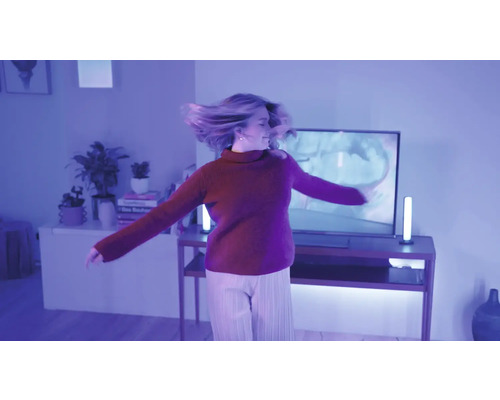 Eine Frau tanzt in einem modern eingerichteten Wohnzimmer das durch violette smarte Lichtleisten neben dem Fernseher beleuchtet wird.