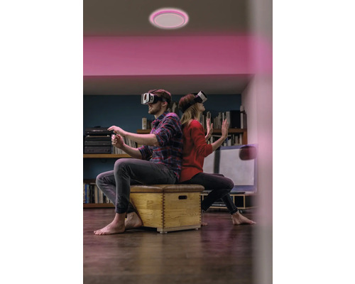 Zwei Personen mit VR-Brillen sitzen unter einer runden LED-Deckenleuchte mit pinkem Licht in einem Wohnzimmer.