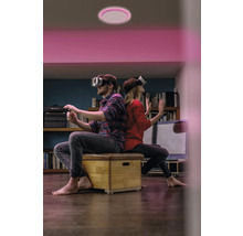 Zwei Personen mit VR-Brillen sitzen unter einer runden LED-Deckenleuchte mit pinkem Licht in einem Wohnzimmer.