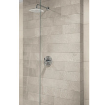 Moderne Dusche mit quadratischem Duschkopf, runder Mischbatterie und hellen Wandfliesen in Steinoptik hinter einer Glastrennwand.
