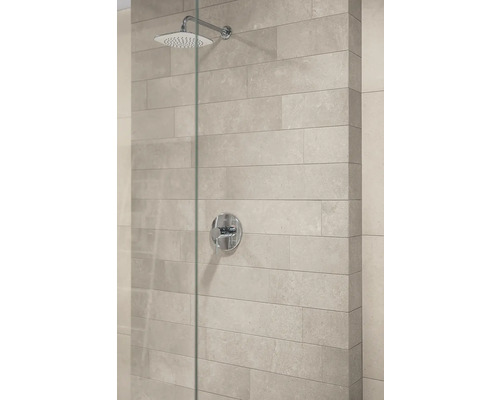 Moderne Dusche mit quadratischem Duschkopf, runder Mischbatterie und hellen Wandfliesen in Steinoptik hinter einer Glastrennwand.