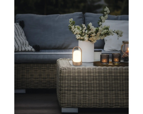 Tragbare LED-Laterne auf einem Couchtisch aus Geflecht in einem Garten-Loungebereich mit Sofa, Kissen und einer Blumenvase.