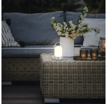 Tragbare, warm beleuchtete LED-Tischleuchte auf einem Rattantisch in einer modern gestalteten Gartenlounge mit Sofa und Blumen.