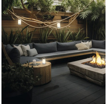 Moderner Garten-Loungebereich mit grauem Sofa, Kissen, einer Feuerstelle, einem Beistelltisch aus Holz mit Laterne und atmosphärischer Beleuchtung.