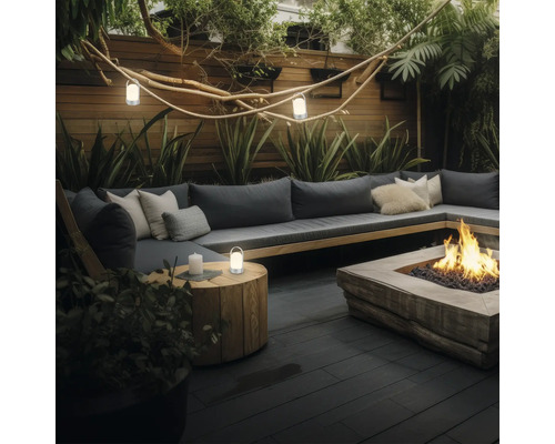 Moderner Garten-Loungebereich mit grauem Sofa, Kissen, einer Feuerstelle, einem Beistelltisch aus Holz mit Laterne und atmosphärischer Beleuchtung.