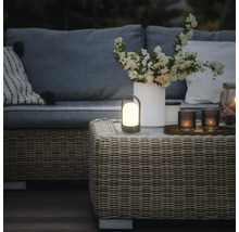 Tragbare LED-Tischleuchte für draußen auf einem Rattantisch in einer warm beleuchteten Gartenlounge mit Sofa, Kissen und einer Blumenvase.