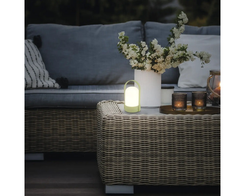 Tragbare LED-Tischleuchte auf einem geflochtenen Gartentisch in einem gemütlich beleuchteten Außenbereich mit Sofa und Blumen.