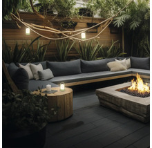 Moderner Außenbereich mit Sofa in L-Form, Feuerstelle aus Holz und Laternen auf dunklen Dielen, umgeben von Pflanzen und einem Holzzaun.