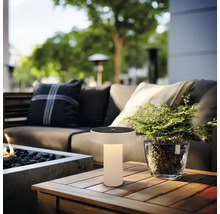 Moderne Solar-Tischleuchte für draußen mit warmem Licht auf einem Holztisch in einem Loungebereich mit Sofa und Pflanze.