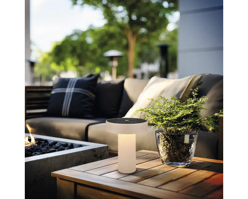 Moderne Solar-Tischleuchte für draußen mit warmem Licht auf einem Holztisch in einem Loungebereich mit Sofa und Pflanze.