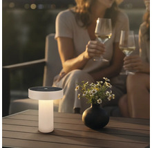 Moderne Solar-Tischleuchte auf einem Holz-Terrassentisch in einer Gartenatmosphäre bei Dämmerung mit Blumen und Personen im Hintergrund.
