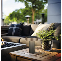 Moderne Tischleuchte auf einem Holztisch auf einer hellen Gartenterrasse mit Sofa, Feuerstelle und Pflanze im Glastopf.