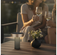 Moderne tragbare Outdoor-Tischleuchte auf einem Holztisch neben einer Blumenvase in gemütlicher Abendstimmung mit Personen im Hintergrund.