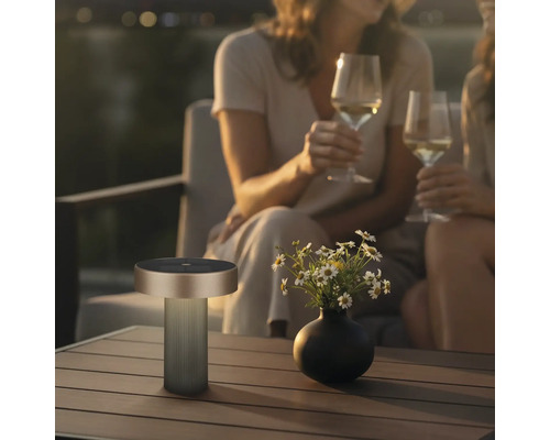 Moderne tragbare Outdoor-Tischleuchte auf einem Holztisch neben einer Blumenvase in gemütlicher Abendstimmung mit Personen im Hintergrund.