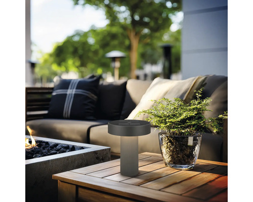 Moderne Außen-Tischleuchte auf einem Holztisch in einer organisierten Garten-Lounge mit Sofa, Pflanze und Feuerstelle.