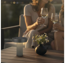 Moderne LED-Solar-Tischleuchte für außen auf einem Holztisch in einer gemütlichen abendlichen Gartenszene mit Personen.