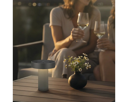 Moderne LED-Solar-Tischleuchte für außen auf einem Holztisch in einer gemütlichen abendlichen Gartenszene mit Personen.