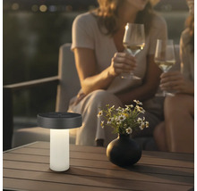Moderne Solar-Tischleuchte auf einem Terrassentisch neben einer Blumenvase, im Hintergrund zwei Personen mit Weingläsern in abendlicher Stimmung.