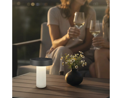 Moderne Solar-Tischleuchte auf einem Terrassentisch neben einer Blumenvase, im Hintergrund zwei Personen mit Weingläsern in abendlicher Stimmung.