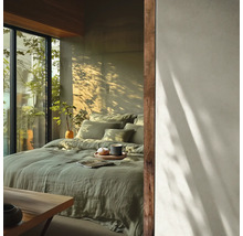 Helles Schlafzimmer mit Bett aus salbeigrüner Leinenbettwäsche, Holzakzenten und großem Fenster mit Blick ins Grüne bei Sonnenlicht.