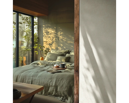 Helles Schlafzimmer mit Bett aus salbeigrüner Leinenbettwäsche, Holzakzenten und großem Fenster mit Blick ins Grüne bei Sonnenlicht.