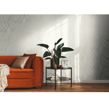 Helle Vliestapete mit grafischem Blattmuster in einem modern eingerichteten Wohnzimmer mit orangefarbenem Sofa, Beistelltisch und Zimmerpflanze.