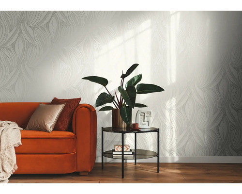 Helle Vliestapete mit grafischem Blattmuster in einem modern eingerichteten Wohnzimmer mit orangefarbenem Sofa, Beistelltisch und Zimmerpflanze.