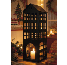 Schwarze Metalllaterne in Hausform mit brennender weißer Kerze in festlicher Weihnachtsumgebung mit Tannenzweigen und warmen Lichterketten.