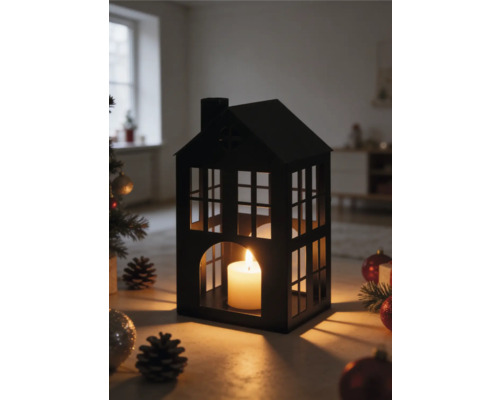 Laterne in Hausform aus schwarzem Metall mit brennender Kerze, umgeben von Weihnachtsdekoration und Tannenzapfen in einem hellen Raum.