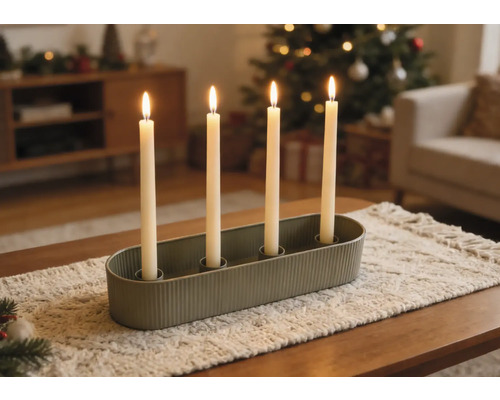 Ovaler grüner Kerzenhalter mit vier brennenden weißen Stabkerzen auf einem Läufer in einem weihnachtlich dekorierten Wohnzimmer.