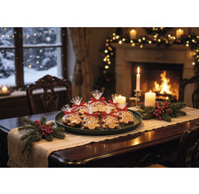 Festlich gedeckter Holztisch mit Lebkuchen in Folienbeuteln, Tannenzweigen, roten Beeren und brennenden Kerzen vor einem warmen Kaminfeuer.