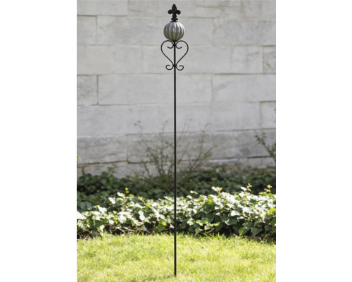 Dekorativer Gartenstab aus Metall mit Keramikkugel, Herz-Element und Lilien-Aufsatz im Garten vor einer Steinmauer.