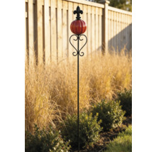 Dekorativer Gartenstecker mit rotem kürbisförmigem Glasornament und schwarzer Lilie an der Spitze in einem Gartenbeet vor einem Holzzaun.