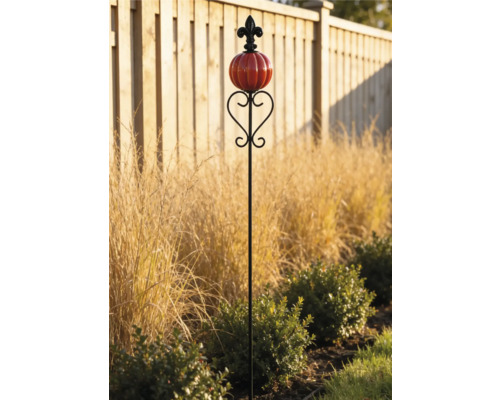 Dekorativer Gartenstecker mit rotem kürbisförmigem Glasornament und schwarzer Lilie an der Spitze in einem Gartenbeet vor einem Holzzaun.