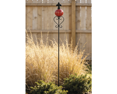 Dekorativer Gartenstab aus Metall mit einer roten Keramikkugel und einer Lilienverzierung in einem Garten mit Ziergras.