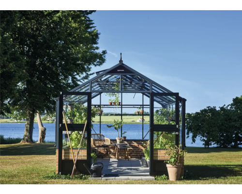 Freistehendes Gewächshaus mit Glaswänden, schwarzem Rahmen und gemauertem Sockel in einem Garten am See.