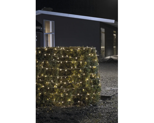 Warmweiß leuchtende LED-Lichterkette für den Außenbereich, um eine Hecke gewickelt, bei Nacht vor einem modernen Haus.