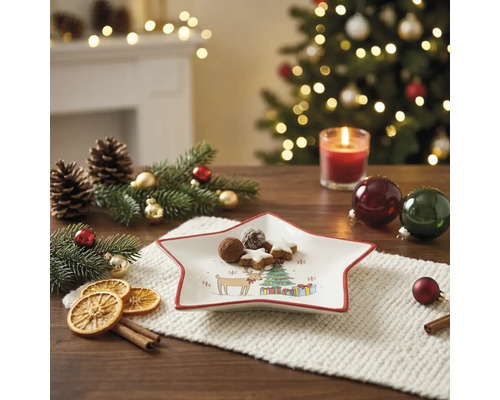 Sternförmiger Weihnachtsteller mit Gebäck auf festlich dekoriertem Tisch mit Tannenzweigen, Zapfen und beleuchtetem Weihnachtsbaum im Hintergrund.