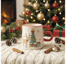 Weihnachtstasse aus Keramik mit Rentier und Tannenbaum auf einer weißen Strickdecke vor einem geschmückten Weihnachtsbaum.