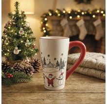 Weiße Keramiktasse mit rotem Henkel, Weihnachtsmann und Schneemann auf Holztisch vor einem beleuchteten Tannenbaum und Kamin.