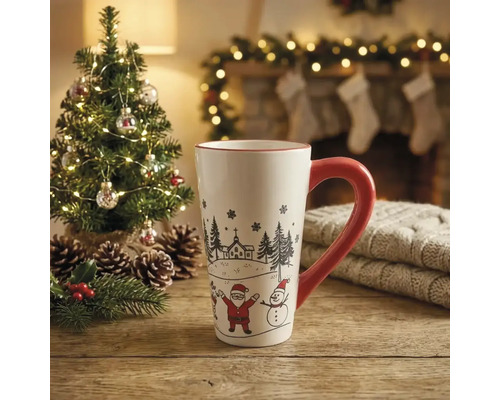 Weiße Keramiktasse mit rotem Henkel, Weihnachtsmann und Schneemann auf Holztisch vor einem beleuchteten Tannenbaum und Kamin.