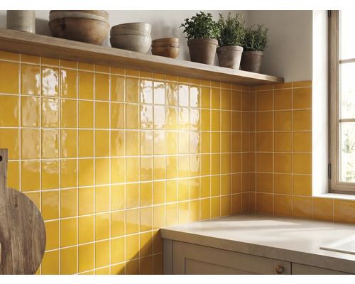 Küchenrückwand mit gelben glänzenden quadratischen Wandfliesen, einem Holzregal mit Schüsseln und Kräutern sowie einer Arbeitsplatte.