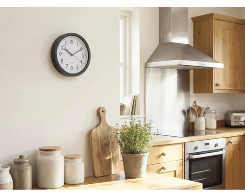 Helle Küche mit einer runden Wanduhr an der Wand, Holzschneidebrett, Kräutertopf und Vorratsdosen auf der Arbeitsplatte.