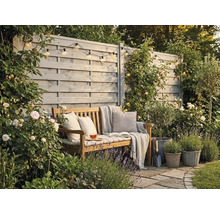Gemütliche Gartenecke mit Holzbank, Kissen und Decke vor einem grauen Sichtschutzzaun aus Holz, umgeben von blühenden Rosen und Lavendel.