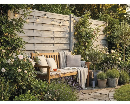 Gemütliche Gartenecke mit Holzbank, Kissen und Decke vor einem grauen Sichtschutzzaun aus Holz, umgeben von blühenden Rosen und Lavendel.