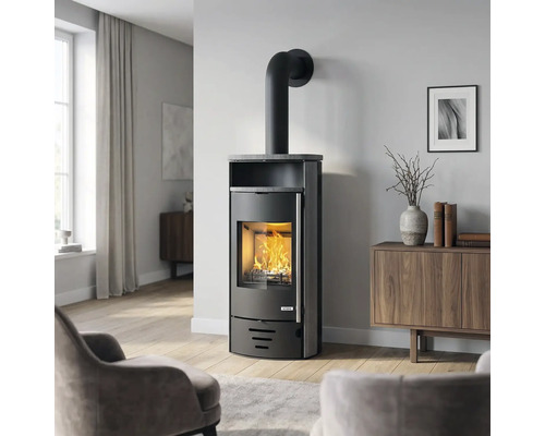 Moderner schwarzer Kaminofen mit ACCENT Logo, brennendem Feuer und Steinplatte in hellem Wohnzimmer mit Holzmöbeln und Parkettboden.