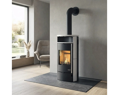 Moderner Kaminofen mit brennendem Holzfeuer in einem hellen Wohnzimmer auf einer Schieferplatte vor einer Wand in Betonoptik.